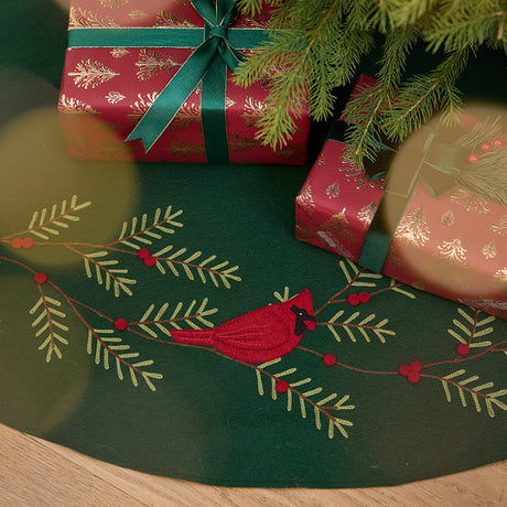 Craftspring’s Woodland Tree Skirt displayed under a Christmas tree with presents — close-up of the embroidered and appliqué cardinal nestled among pine branches and red berries, highlighting festive holiday details, ethically crafted by artisans using sustainable wool.