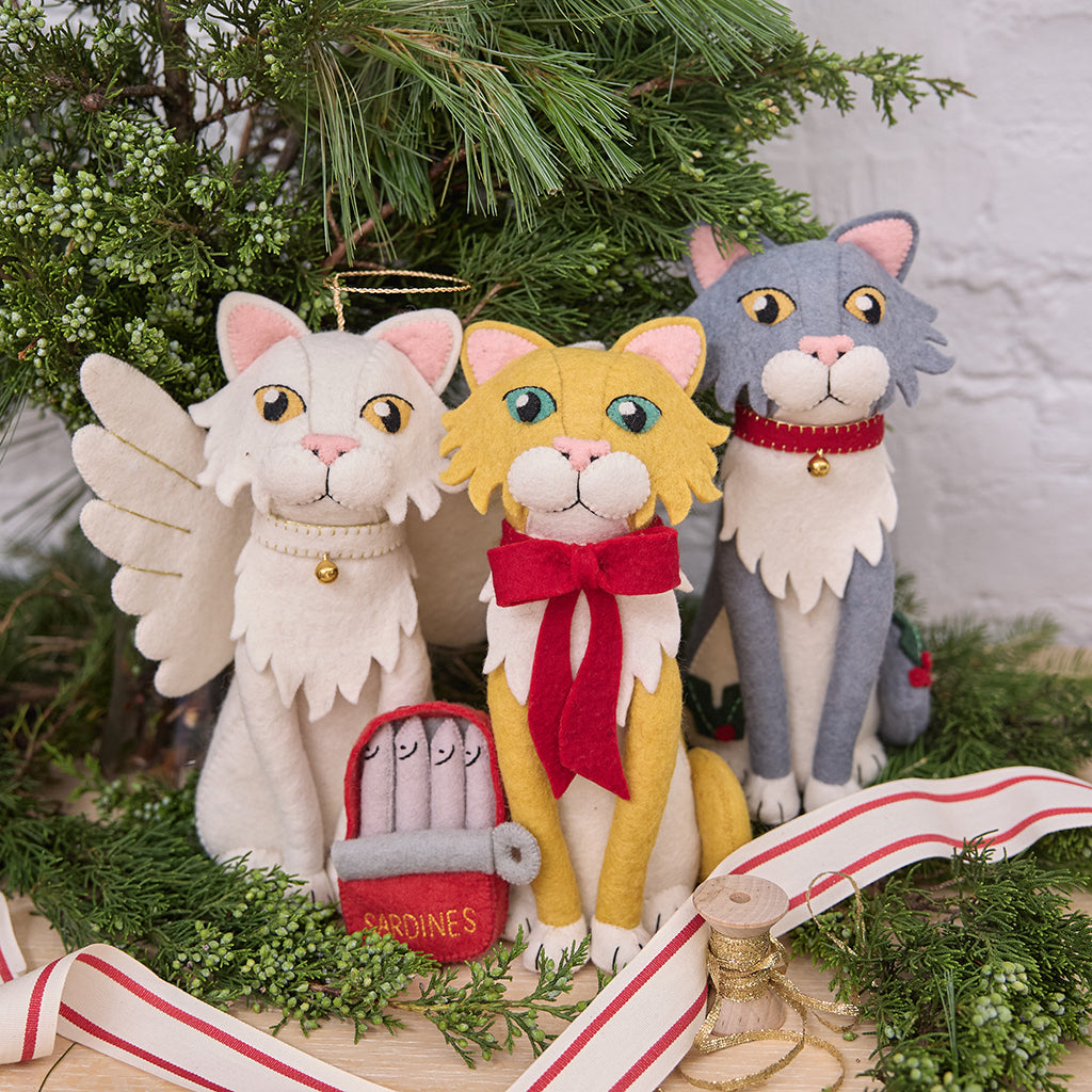 Handmade felt cat tree toppers from Craftspring, featuring a gray cat with holly and a collar with bell, an orange cat with a red bow, and a white angel cat—displayed among fresh evergreens, perfect for festive cat lovers and charming holiday decorating, ethically crafted by artisans using sustainable wool.