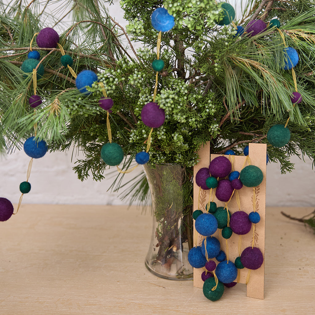Handmade felt Jewel Tone Ball Garland from Craftspring, featuring deep jewel-tone felt balls strung on a gold ribbon—displayed among evergreen branches, perfect for adding rich, festive color and heirloom-quality holiday charm, ethically crafted by artisans using sustainable wool.