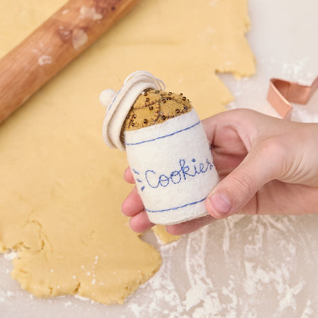 Handmade felt Cookie Jar ornament from Craftspring, featuring chocolate chip cookies with embroidered and beaded details—held in hand in front of rolled out cookie dough, perfect for festive kitchen decor and holiday gifting, ethically crafted by artisans using sustainable wool.