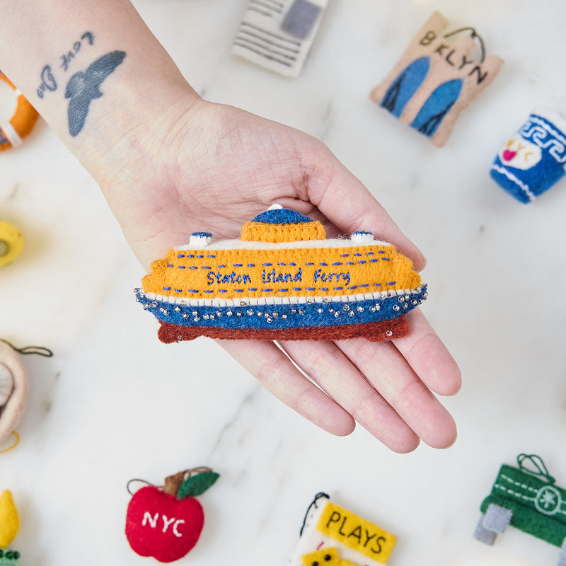 Craftspring’s handmade felt Staten Island Ferry ornament — featuring intricate embroidered details and beaded waves on the iconic orange ferry, held in hand above the NYC Mini Ornaments Set of 24 for a festive holiday scene, perfect for Christmas decorating or gifting, ethically crafted by artisans using sustainable wool.