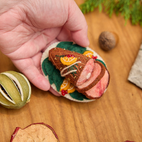 Craftspring’s handmade felt Spiral Ham Platter ornament — a festive ham with intricate beaded and embroidered details, held in a hand above a harvest table for scale, perfect for Thanksgiving decorating or gifting, ethically crafted by artisans using sustainable wool.
