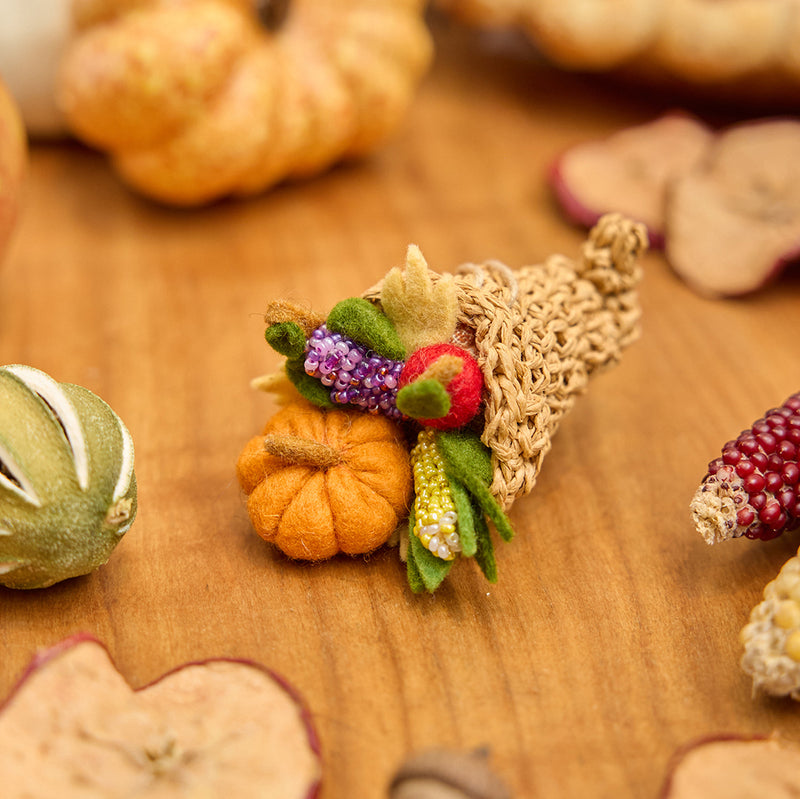 Craftspring’s handmade felt Cornucopia ornament — a crocheted raffia cornucopia filled with felt and beaded vegetables and fruit, displayed on a harvest table with pumpkins and dried apple slices for a cozy holiday scene, perfect for Christmas decorating or gifting, ethically crafted by artisans using sustainable wool.
