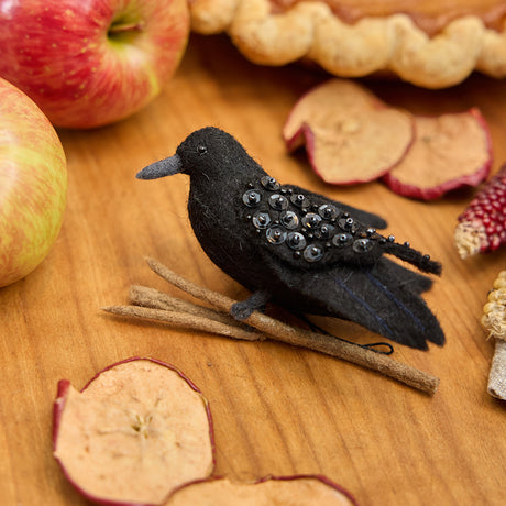 Craftspring’s handmade felt Crow ornament — featuring shimmering beaded wings and perched on a branch, displayed on a harvest table with apples and a pie for a festive autumn scene, perfect for Halloween, Thanksgiving, and holiday gifting, ethically crafted by artisans using sustainable wool.