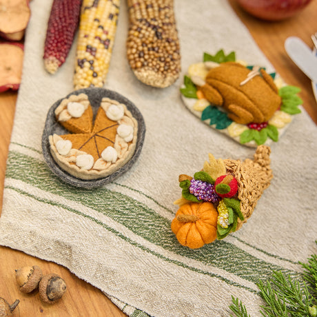 Craftspring’s handmade felt Cornucopia, Turkey Dinner, and Pumpkin Pie ornaments — a crocheted raffia cornucopia filled with felt and beaded vegetables and fruit, a roasted turkey with autumn sides, and a plump pumpkin pie, displayed on a harvest table with autumn corn for a festive Thanksgiving scene, perfect for holiday decorating or gifting, ethically crafted by artisans using sustainable wool.