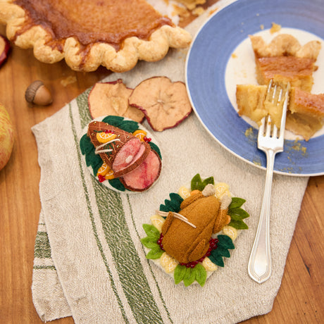 Craftspring’s handmade felt Spiral Ham Platter and Turkey Dinner ornaments — a festive ham with intricate beaded and embroidered details and a roasted turkey with autumn sides, displayed on a harvest table with pumpkin pie for a cozy holiday scene, perfect for Thanksgiving decorating or gifting, ethically crafted by artisans using sustainable wool.