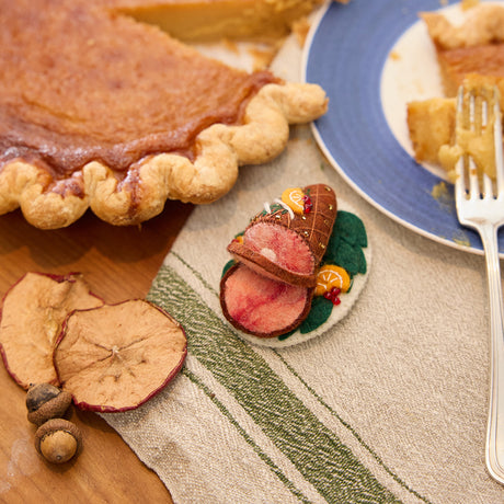 Craftspring’s handmade felt Spiral Ham Platter ornament — a festive ham with intricate beaded and embroidered details, displayed on a harvest table with pumpkin pie for a cozy holiday scene, perfect for Thanksgiving decorating or gifting, ethically crafted by artisans using sustainable wool.