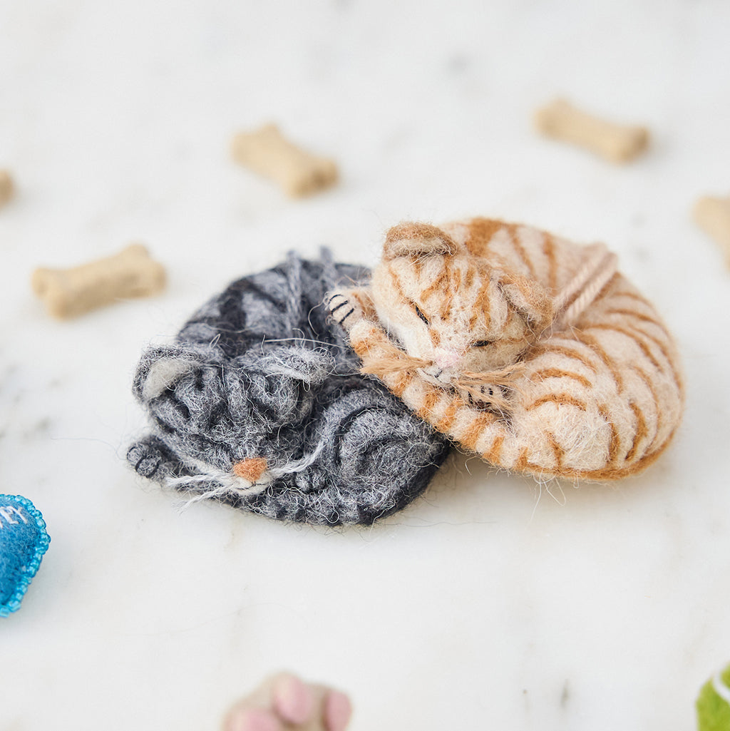 Craftspring’s needle-felted Sleepy Orange Cat and Sleepy Gray Cat ornaments displayed among pet treats and toys — both with cute faces, whiskers, and charming details, creating a playful and festive holiday scene, perfect for gifting, ethically crafted by artisans using sustainable wool.