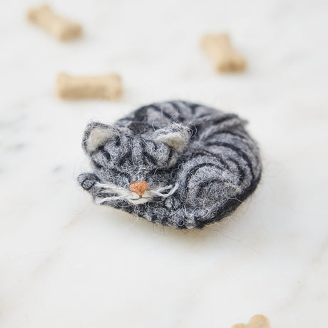 Craftspring’s needle-felted Sleepy Gray Cat Ornament displayed among pet treats and toys — with whiskers and a cute face, creating a playful and festive holiday scene, perfect for gifting, ethically crafted by artisans using sustainable wool.