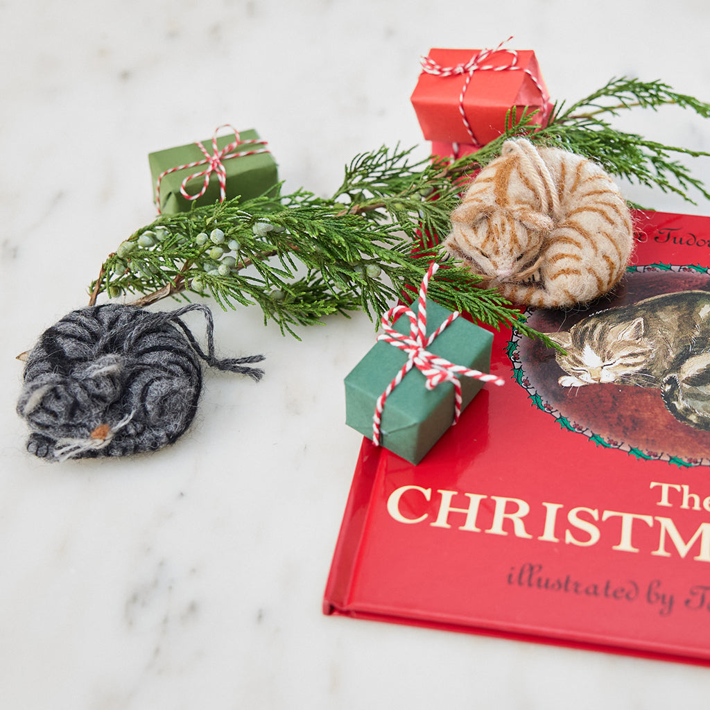 Craftspring’s needle-felted Sleepy Orange Cat and Sleepy Gray Cat ornaments displayed with The Christmas Cat book, evergreens, and mini presents — both with cute faces, whiskers, and charming details, creating a cozy and festive holiday scene, perfect for gifting, ethically crafted by artisans using sustainable wool.