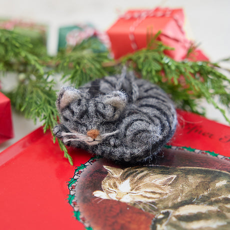 Craftspring’s needle-felted Sleepy Gray Cat Ornament displayed on The Christmas Cat book with evergreens and mini presents — with whiskers and a cute face, creating a warm and festive holiday scene, perfect for gifting, ethically crafted by artisans using sustainable wool.