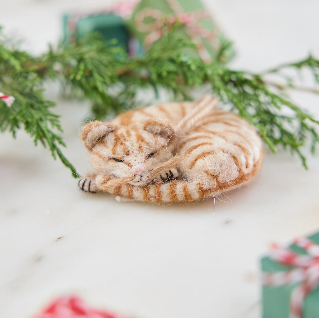 Craftspring’s needle-felted Sleepy Orange Cat Ornament, a striped tabby with whiskers and a cute face, displayed with evergreens and mini presents — a cozy and festive holiday scene, perfect for gifting, ethically crafted by artisans using sustainable wool.
