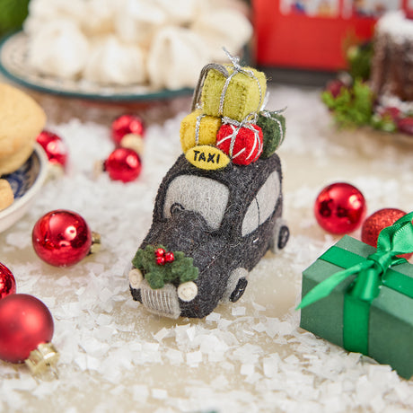 Craftspring’s handmade felt London Black Cab ornament — featuring beaded and embroidered details with festive presents on top of the car and holly accents, displayed in snow with Christmas baubles and treats for a joyful holiday scene, perfect for Christmas decorating or gifting, ethically crafted by artisans using sustainable wool.