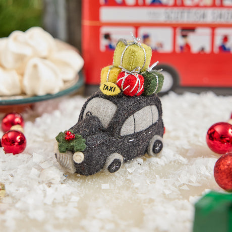 Craftspring’s handmade felt London Black Cab ornament — featuring beaded and embroidered details with festive presents on top of the car and holly accents, displayed in snow with Christmas baubles and treats for a joyful holiday scene, perfect for Christmas decorating or gifting, ethically crafted by artisans using sustainable wool.