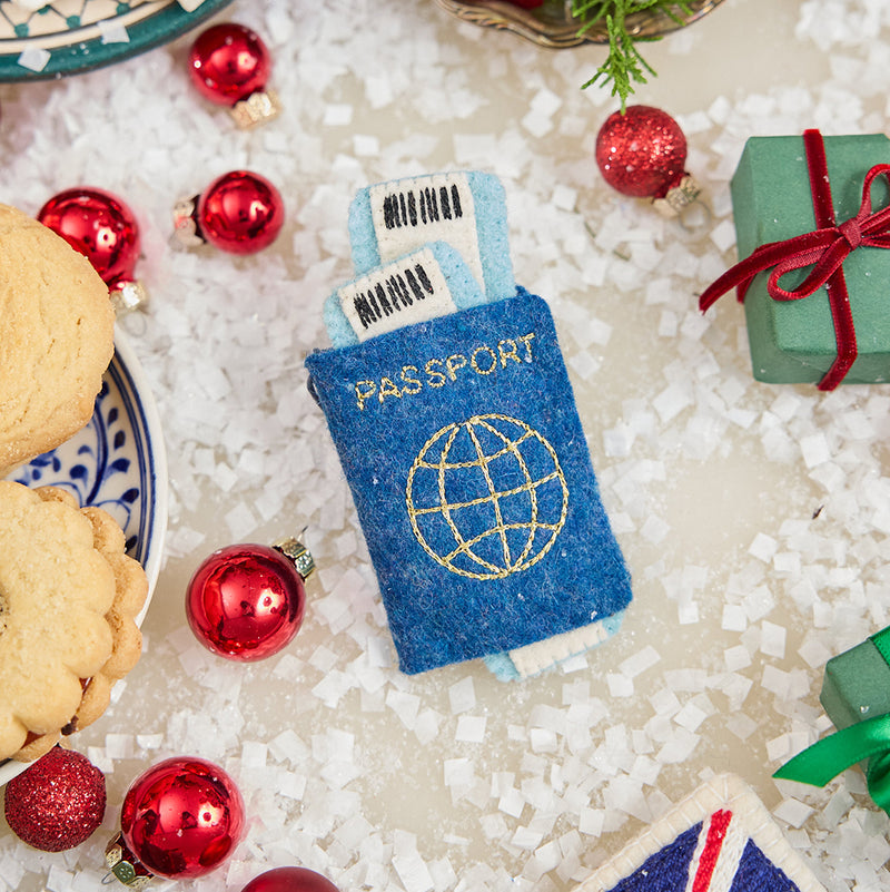 Craftspring’s handmade felt Passport ornament — featuring applique and embroidered details with two plane tickets tucked inside and passport stamps on the back, displayed with Christmas baubles and mini presents for a festive holiday scene, perfect for Christmas decorating or gifting, ethically crafted by artisans using sustainable wool.