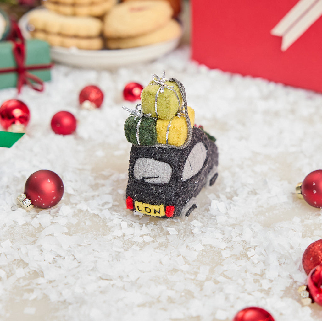 Craftspring’s handmade felt London Black Cab ornament — featuring beaded and embroidered details with festive presents on top of the car and holly accents, displayed in snow with Christmas baubles and treats for a joyful holiday scene, perfect for Christmas decorating or gifting, ethically crafted by artisans using sustainable wool.