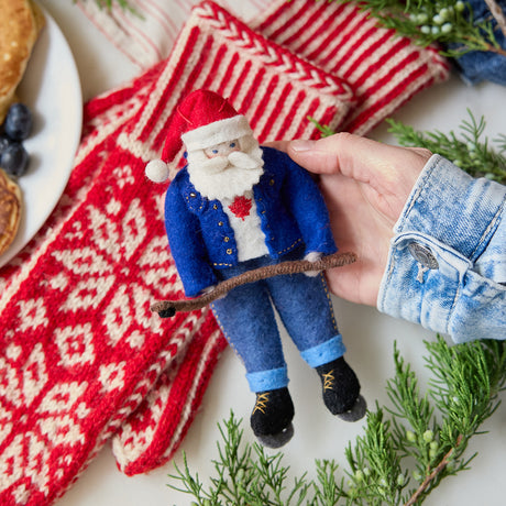 Craftspring’s Canadian Santa Ornament — felt Santa wearing a denim jacket and jeans with ice skates, holding a hockey stick, and featuring a Canadian flag maple leaf — held in hand over mittens and evergreens, capturing playful holiday charm, festive warmth, and heirloom-quality gifting, ethically crafted by artisans using sustainable wool.