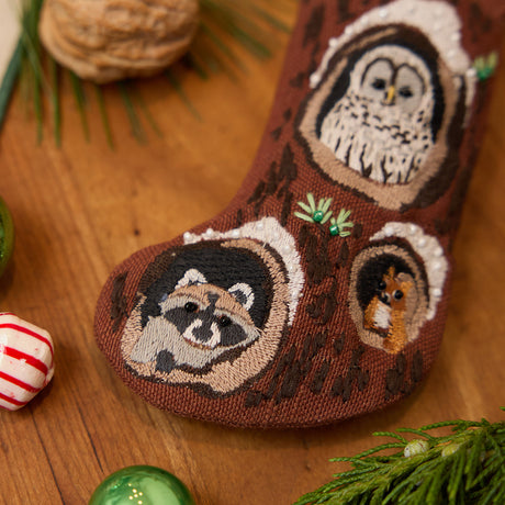 Craftspring’s handmade linen Woodland Mini Stocking ornament — featuring intricate beaded and embroidered details with an owl, raccoon, and squirrel peeking out of holes in a tree, displayed with evergreens, Christmas baubles, and peppermint candies for a festive holiday scene, perfect for Christmas decorating or gifting, ethically crafted by artisans using sustainable materials.