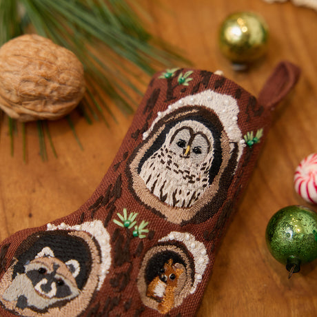Craftspring’s handmade linen Woodland Mini Stocking ornament — featuring intricate beaded and embroidered details with an owl, raccoon, and squirrel peeking out of holes in a tree, displayed with evergreens, Christmas baubles, and peppermint candies for a festive holiday scene, perfect for Christmas decorating or gifting, ethically crafted by artisans using sustainable materials.