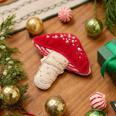 Craftspring’s handmade satin and velvet Mushroom Beaded Ornament — featuring intricate beaded and embroidered details with a gnome peeking out of a door on one side of the mushroom, displayed with Christmas baubles, peppermint candies, and mini presents for a festive holiday scene, perfect for Christmas decorating or gifting, ethically crafted by artisans using sustainable materials.