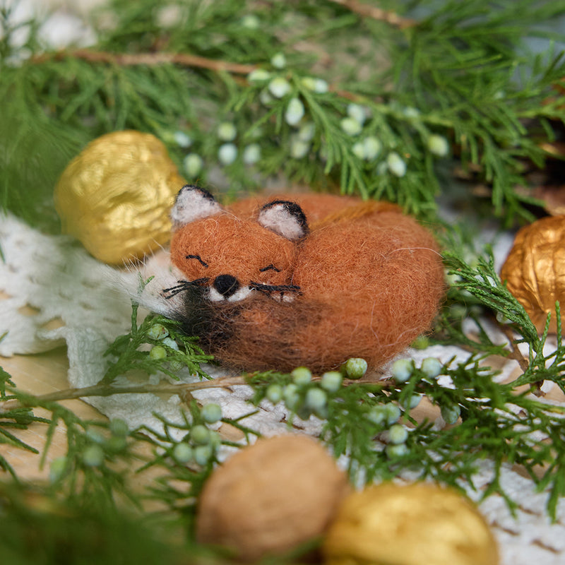 Craftspring’s Sleepy Fox Ornament — handmade with a curled-up body, cute face, fluffy tail, and whiskers — nestled among evergreens with walnuts, capturing the warmth, charm, and heirloom-quality beauty of holiday gifting, ethically crafted by artisans using sustainable wool.