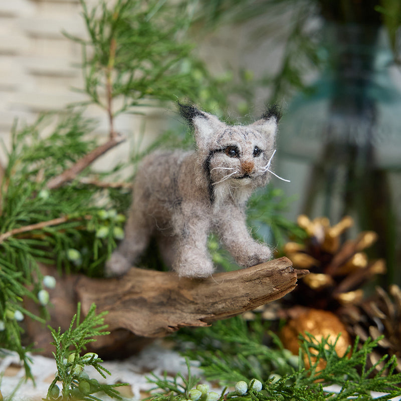 Craftspring’s Lynx ornament displayed standing on a stick among evergreens and pinecones — handmade from sustainable wool with intricate needle-felted details, whiskers, and ear tufts, ethically crafted by artisans for Christmas decorating and gifting.