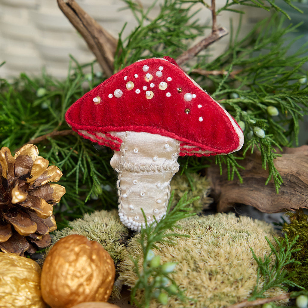 Craftspring’s handmade satin and velvet Mushroom Beaded Ornament — featuring intricate beaded and embroidered details with a gnome peeking out of a door on one side of the mushroom, displayed nestled among moss, evergreens, and golden walnuts for a cozy woodland holiday scene, perfect for Christmas decorating or gifting, ethically crafted by artisans using sustainable materials.