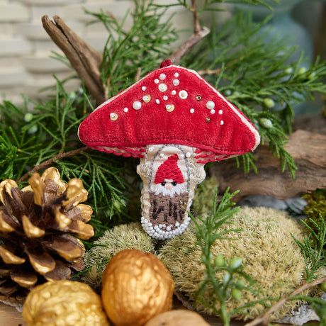 Craftspring’s handmade satin and velvet Mushroom Beaded Ornament — featuring intricate beaded and embroidered details with a gnome peeking out of a door on one side of the mushroom, displayed nestled among moss, evergreens, and golden walnuts for a cozy woodland holiday scene, perfect for Christmas decorating or gifting, ethically crafted by artisans using sustainable materials.