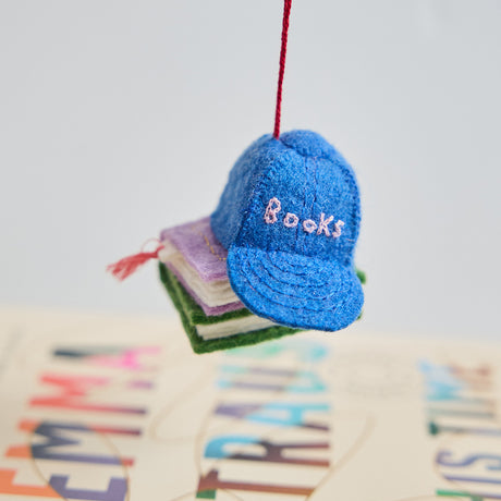 Lifestyle shot of Craftspring’s Books Are Magic Hat with Books ornament, created in collaboration with the Books Are Magic bookstore—whimsical blue hat on a stack of books, hanging above a book for a festive holiday touch.