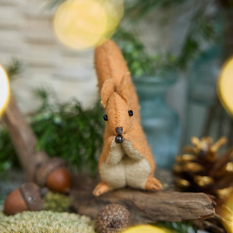 Craftspring’s handmade felt Red Squirrel ornament — featuring delicate beaded and embroidered details, displayed sitting on a stick surrounded by acorns, pinecones, and moss for a cozy woodland holiday scene, perfect for Christmas decorating or gifting, ethically crafted by artisans using sustainable wool.