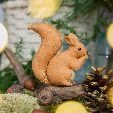 Craftspring’s handmade felt Red Squirrel ornament — featuring delicate beaded and embroidered details, displayed sitting on a stick surrounded by acorns, pinecones, and moss for a cozy woodland holiday scene, perfect for Christmas decorating or gifting, ethically crafted by artisans using sustainable wool.