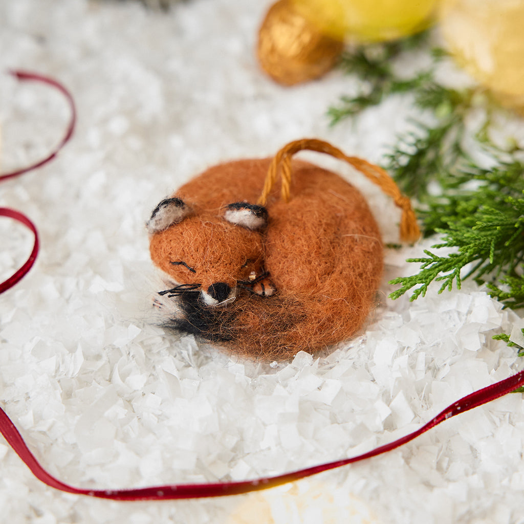 Craftspring’s Sleepy Fox Ornament — needle felted, curled up with a cute face, fluffy tail, and whiskers — displayed on snow among evergreens, evoking cozy holiday magic, woodland charm, and the joy of Christmas gifting, ethically crafted by artisans using sustainable wool.