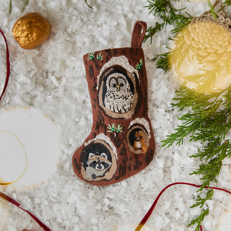 Craftspring’s handmade linen Woodland Mini Stocking ornament — featuring intricate beaded and embroidered details with an owl, raccoon, and squirrel peeking out of holes in a tree, displayed with evergreens and golden walnuts for a festive holiday scene, perfect for Christmas decorating or gifting, ethically crafted by artisans using sustainable materials.