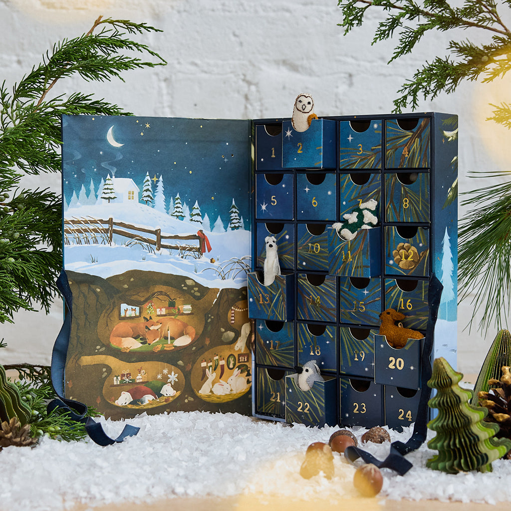 Craftspring’s handmade Woodland Minis Advent Calendar — showing the inside with 24 drawers and mini felt woodland ornaments peeking out, alongside an illustration of woodland animals nestled in cozy holiday burrows, perfect for Christmas decorating or gifting, ethically crafted by artisans using sustainable wool.