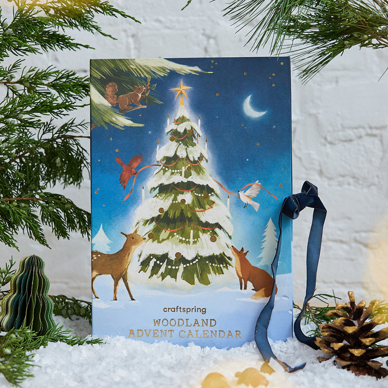 Craftspring’s handmade Woodland Minis Advent Calendar — showing the front illustration of animals surrounding a Christmas tree, displayed with evergreens and snow for a festive woodland holiday scene, perfect for Christmas decorating or gifting, ethically crafted by artisans using sustainable wool.