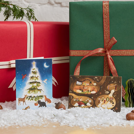 Craftspring’s Woodland Tree and Woodland Burrows Greeting Cards — displayed on snow in front of a present, featuring woodland animals around a glowing Christmas tree and animals nestled in cozy holiday burrows, ethically printed in the USA on natural textured paper, perfect for sharing holiday cheer and gifting.