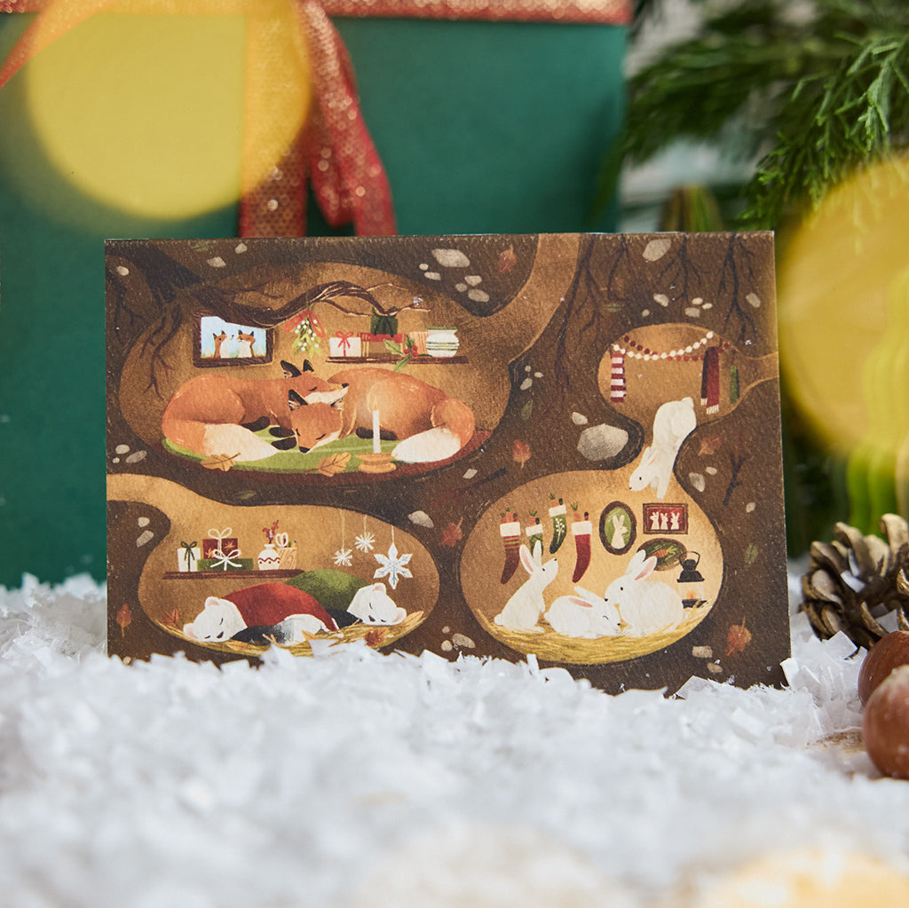 Craftspring’s Woodland Burrows Greeting Card — displayed on snow in front of a present, featuring woodland animals nestled in cozy holiday burrows, ethically printed in the USA on natural textured paper, perfect for sharing holiday cheer and gifting.
