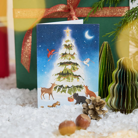 Craftspring’s Woodland Tree Greeting Card — displayed on snow in front of a present, featuring woodland animals around a glowing winter Christmas tree, ethically printed in the USA on natural textured paper, perfect for sharing holiday cheer and gifting.