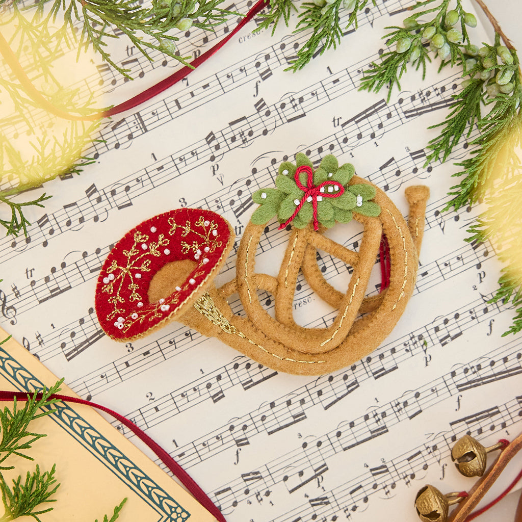 Craftspring’s French Horn Ornament displayed on sheet music with fresh evergreens — handmade felt holiday decor with intricate golden embroidery, beading, and mistletoe, ethically crafted by artisans using sustainable wool.