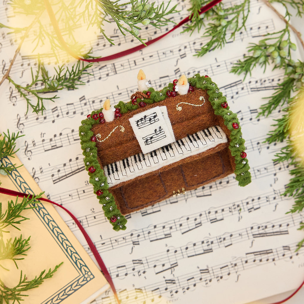 Craftspring’s Christmas Piano Ornament displayed on sheet music with evergreens — handmade felt holiday decor with intricate beaded and embroidered details, heirloom-quality ornament ethically crafted by artisans using sustainable wool.