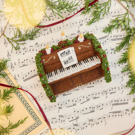 Craftspring’s Christmas Piano Ornament displayed on sheet music with evergreens — handmade felt holiday decor with intricate beaded and embroidered details, heirloom-quality ornament ethically crafted by artisans using sustainable wool.