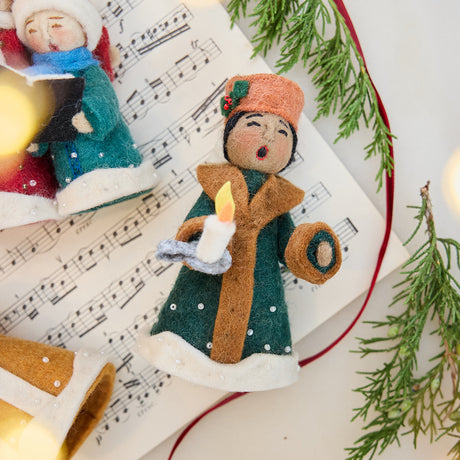 Caroler Holding a Candle Ornament displayed on sheet music with fresh evergreens — handmade felt ornament with intricate applique and embroidered snow, holding a candle, ethically crafted by artisans using sustainable wool.