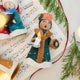 Caroler Holding a Candle Ornament displayed on sheet music with fresh evergreens — handmade felt ornament with intricate applique and embroidered snow, holding a candle, ethically crafted by artisans using sustainable wool.