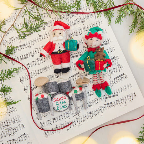 Craftspring’s Christmas Drum Set Ornament, Elf with Gifts Ornament, and Little Santa with Gift styled together on sheet music with fresh evergreens — handmade felt Christmas ornaments with intricate embroidery and festive details, heirloom-quality holiday decor ethically crafted by artisans using sustainable wool.