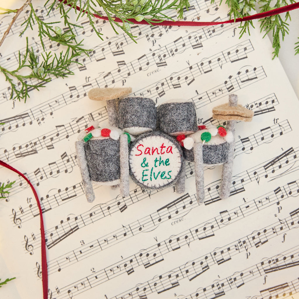Craftspring’s Christmas Drum Set Ornament styled on sheet music with fresh evergreens — handmade felt holiday decor with embroidered “Santa and the Elves” band name and festive garland, ethically crafted by artisans using sustainable wool.