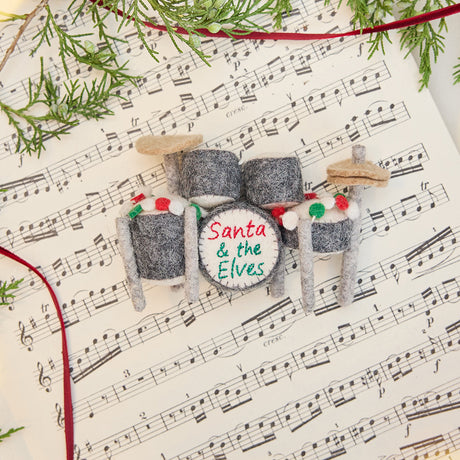 Craftspring’s Christmas Drum Set Ornament styled on sheet music with fresh evergreens — handmade felt holiday decor with embroidered “Santa and the Elves” band name and festive garland, ethically crafted by artisans using sustainable wool.
