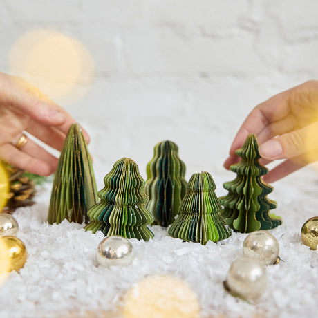 Craftspring’s Green Honeycomb Paper Mini Trees — Set of 5, displayed on snow with Christmas bobbles, with hands reaching in to arrange them — highlighting their shades of green, gold sparkle, and varying shapes, handmade by artisans from recycled cotton waste for a playful holiday scene.