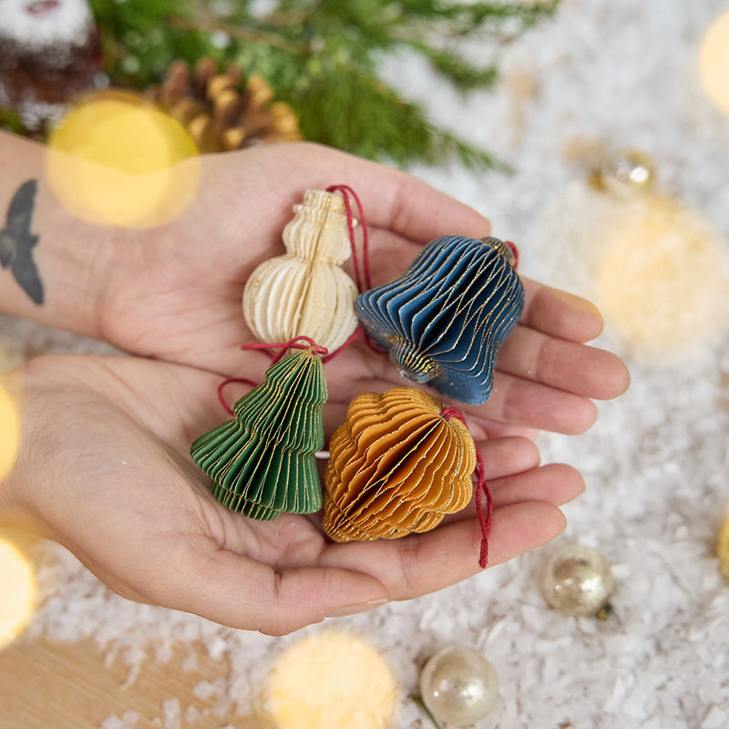 Craftspring’s Classic Christmas Honeycomb Paper Mini Ornaments — Set of 4, held in hand above snow and evergreens, featuring the snowman, bell, pinecone, and tree with gold sparkly edges, handmade by artisans from recycled cotton waste for playful holiday gifting charm.