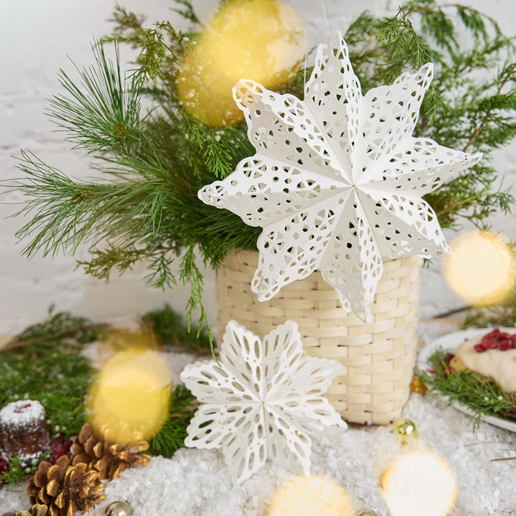 Craftspring’s Paper Snowflakes Christmas Ornaments — Set of 2, large white snowflakes with intricate cutout patterns, one slightly larger than the other — displayed on snow in front of a basket of evergreens, creating a festive holiday scene, ethically handmade by artisans from recycled cotton waste.