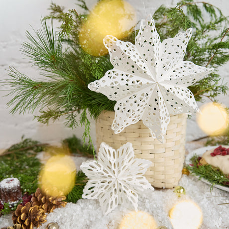 Craftspring’s Paper Snowflakes Christmas Ornaments — Set of 2, large white snowflakes with intricate cutout patterns, one slightly larger than the other — displayed on snow in front of a basket of evergreens, creating a festive holiday scene, ethically handmade by artisans from recycled cotton waste.